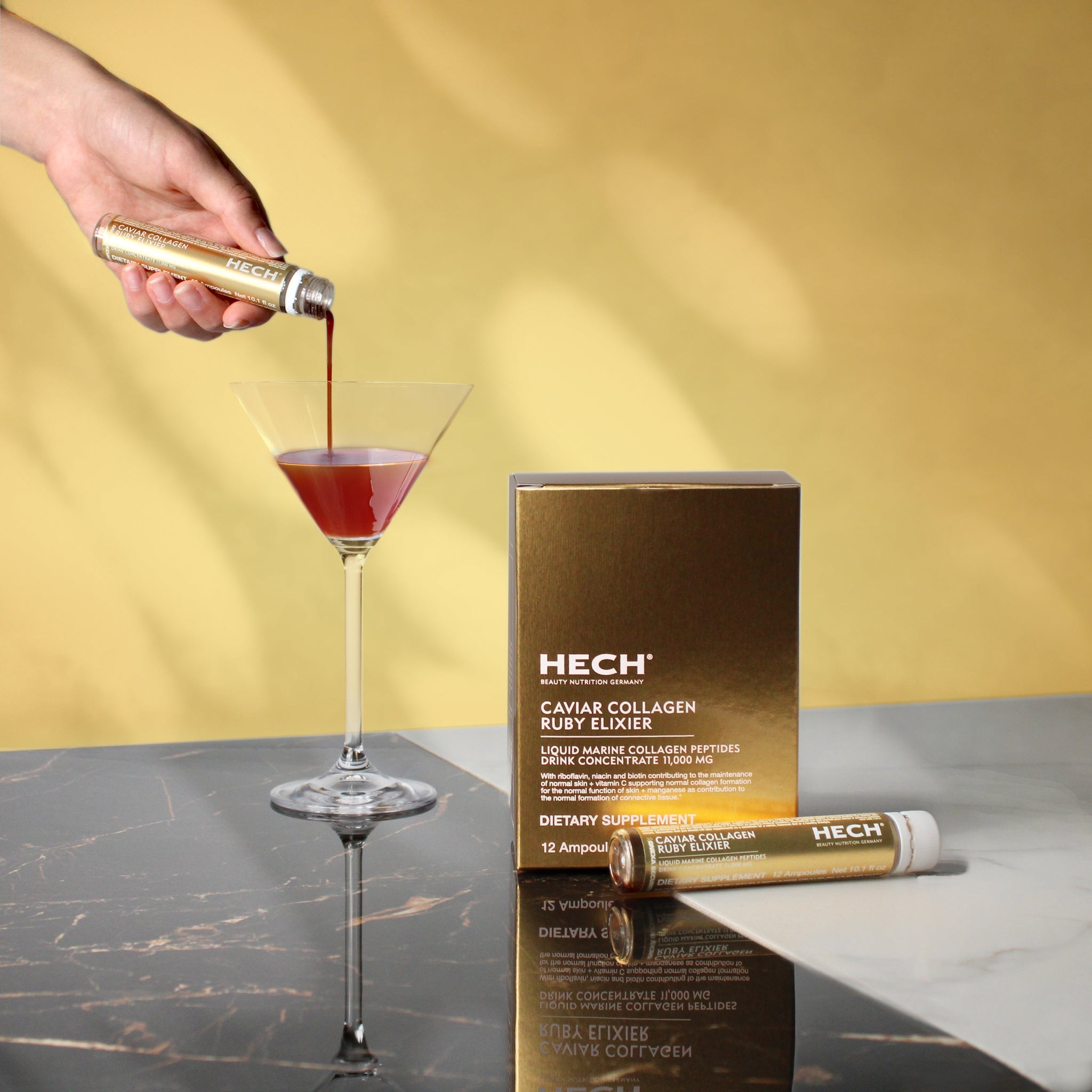 Hand pouring a red liquid into a martini glass with HECH Caviar Collagen Elixir packaging in the background.